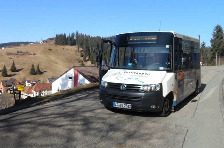 Ein weißer Kleinbus fährt auf einer offenen Landstraße. Man sieht der Bus mittels einer schrägen Vorderansicht. Es ist ein sonniger Tag mit blauem Himmel.